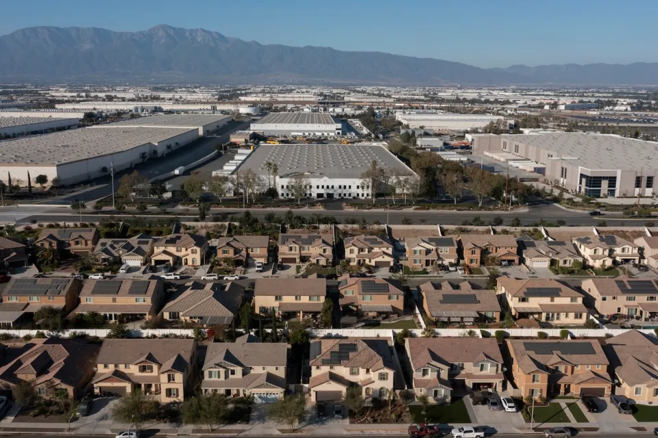 Aerial view of large warehouses across from development of suburban single-family homes in Jurupa, California with desert mountains in background.